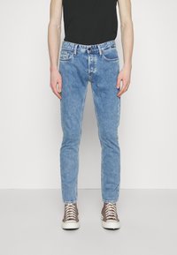 Světle modré džíny s úzkým střihem, designem s pěti kapsami a kontrastním šitím. Nosené s černou košilí a hnědými teniskami.