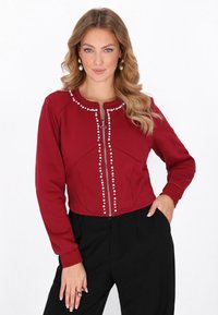 Chaqueta corta roja con cremallera frontal, cuello redondo y detalles decorativos que parecen perlas a lo largo del cierre. Tela suave y diseño ajustado.
