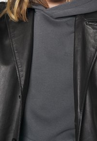 Giubbotto di pelle nera sopra un hoodie grigio. L'hoodie ha una consistenza morbida, con un grande cappuccio e un marchio discreto sul petto.