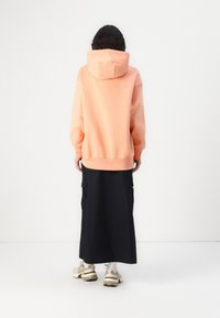 Sweat à capuche pêche avec une grande capuche, coupe décontractée, poignets côtelés, associé à une longue jupe noire et des baskets beiges. Design minimaliste et look sans couture.