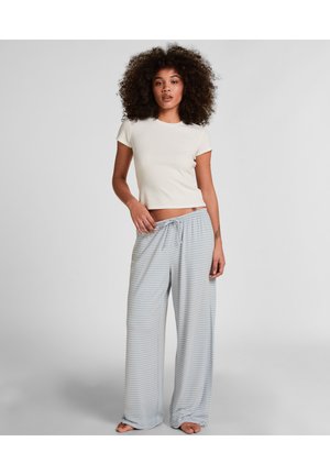 Vrouw met krullend haar, gekleed in een wit crop top met korte mouwen en losse, blauw-wit gestreepte broek met trekkoord, staand op blote voeten op een grijze vloer.