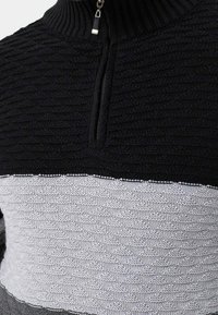 Knitted pullover with a high collar, featuring a textured black upper section and a patterned gray and white lower section. Zipper detail present.