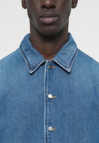 Ανοιχτό μπλε τζιν μπουφάν με μυτερό γιακά, κούμπωμα με κουμπιά μπροστά και εμφανή ραφές. Διαθέτει δύο κουμπιά με διακοσμητικές κλωστές.