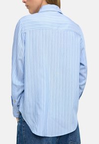 Jasnoniebieska koszula w paski z długim rękawem, zapięciem na guziki przy mankietach oraz lekko zaokrąglonym dołem. Tkanina ma miękką teksturę oraz subtelne pionowe linie.