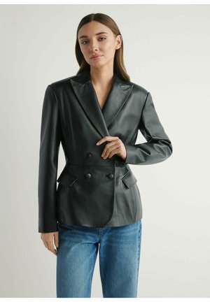 Kobieta ubrana w czarną, dwurzędową skórzaną marynarkę i niebieskie dżinsy, pozująca z jedną ręką regulującą klapę marynarki na jednolitym tle.