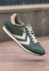Scarpa sportiva verde con accenti crema, tessuto testurizzato, design a chevron bianco, costruzione leggera e suola in gomma piatta.
