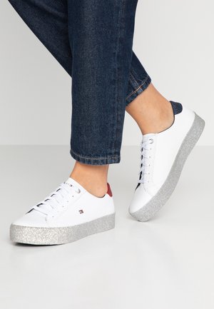 Personne portant des baskets blanches avec des semelles argentées pailletées et un jean bleu foncé sur un fond gris clair uni.
