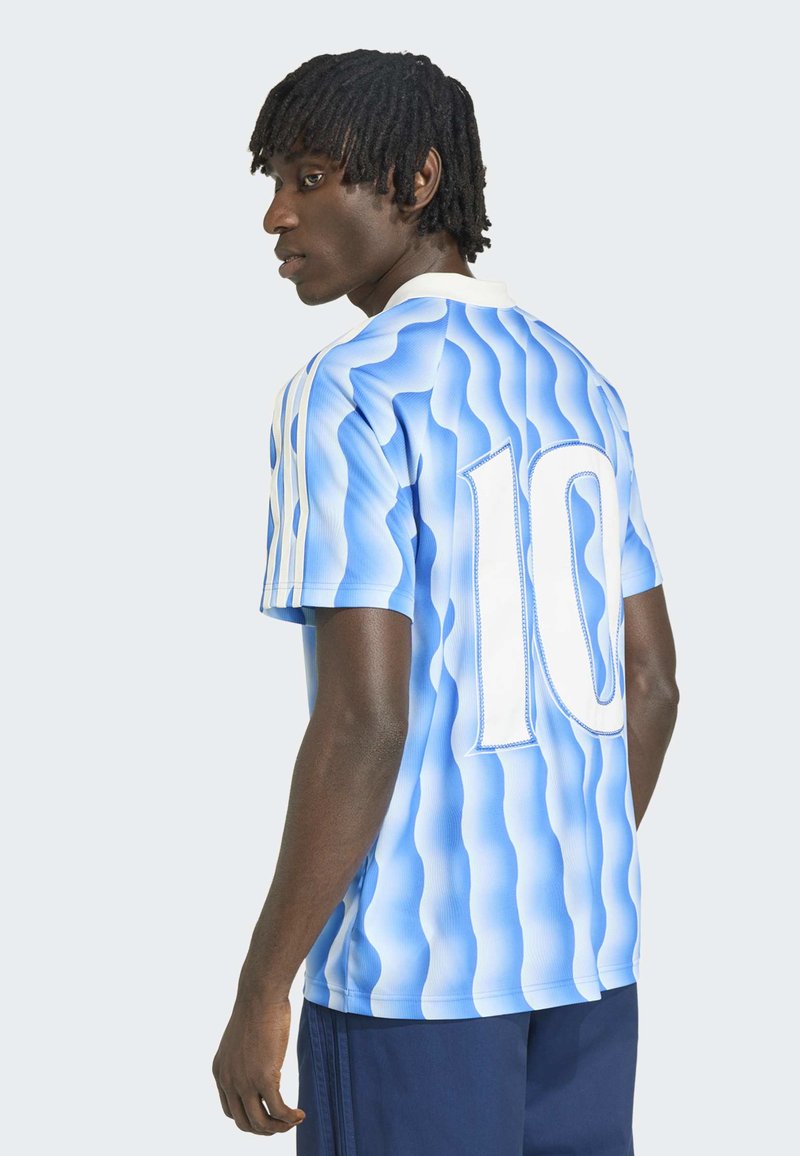 Junger Mann mit dunkler Haut und Dreadlocks, der ein welliges blau-weiß gestreiftes Sporttrikot mit der Nummer 10 auf dem Rücken und dunkle Shorts trägt.