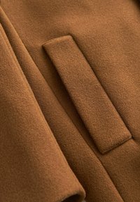 Gros plan sur la poche à rabat d'un manteau en laine marron, mettant en avant le tissu doux et texturé ainsi que les coutures précises.