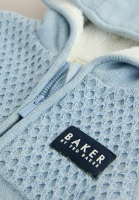 Lysblå strikket zip-jakke med et struktureret mønster, hvid fleeceforing og et mørkeblåt stofmærke med teksten "BAKER by TED BAKER."