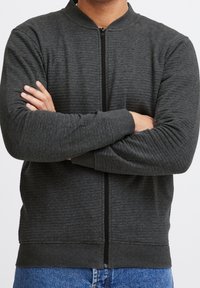 Czarna szara kurtka na zamek błyskawiczny z teksturowanym wzorem, ściągaczami przy rękawach i stójką, noszona na niebieskich dżinsach, z założonymi rękami.