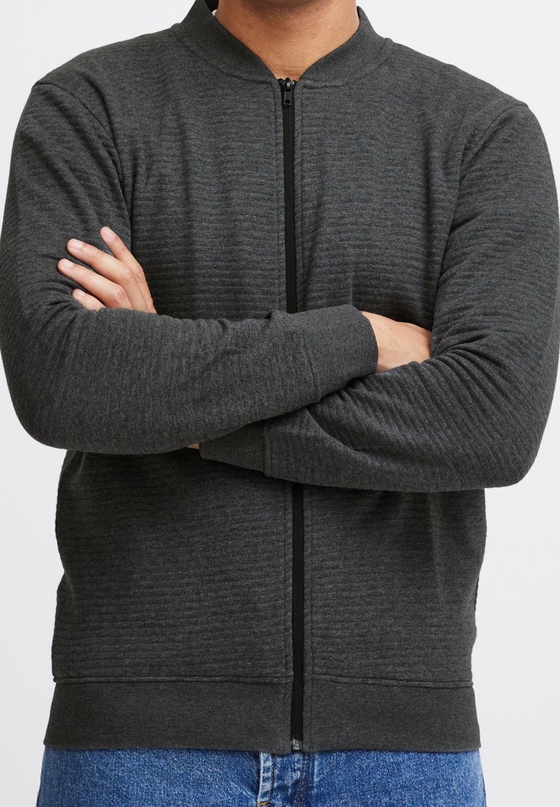 Czarna szara kurtka na zamek błyskawiczny z teksturowanym wzorem, ściągaczami przy rękawach i stójką, noszona na niebieskich dżinsach, z założonymi rękami.