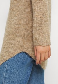 Pull beige en maille à la coupe ample, présentant une texture côtelée et un ourlet fendu. Associé à un jean denim bleu clair.