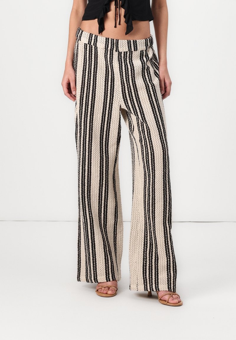 Femme portant un pantalon crème taille haute à jambes larges avec des rayures verticales noires et des sandales, debout devant un fond blanc uni.