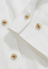 Cămașă albă dintr-un material din bumbac neted, cu guler tip bandă și nasturi bej cu un model marmorat pe marginea și manșetele mânecilor.