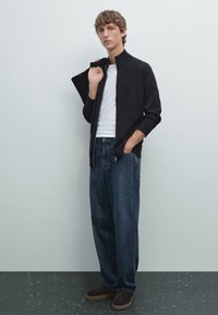 Schwarze Zip-Jacke über einem weißen T-Shirt, kombiniert mit locker sitzenden blauen Jeans. Dunkelbraune Schuhe runden das Outfit vor einem neutralen Hintergrund ab.