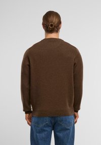 Brauner Strickpullover mit rundem Halsausschnitt und langen Ärmeln, der eine strukturierte Oberfläche aufweist. Kombiniert mit blauen Jeans.