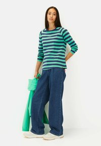 Gestreepte langemouwen shirt in groen en blauw, gecombineerd met losse blauwe spijkerbroeken. Het model houdt een lichtgroene jas vast. Neutrale sneakers maken de look compleet.