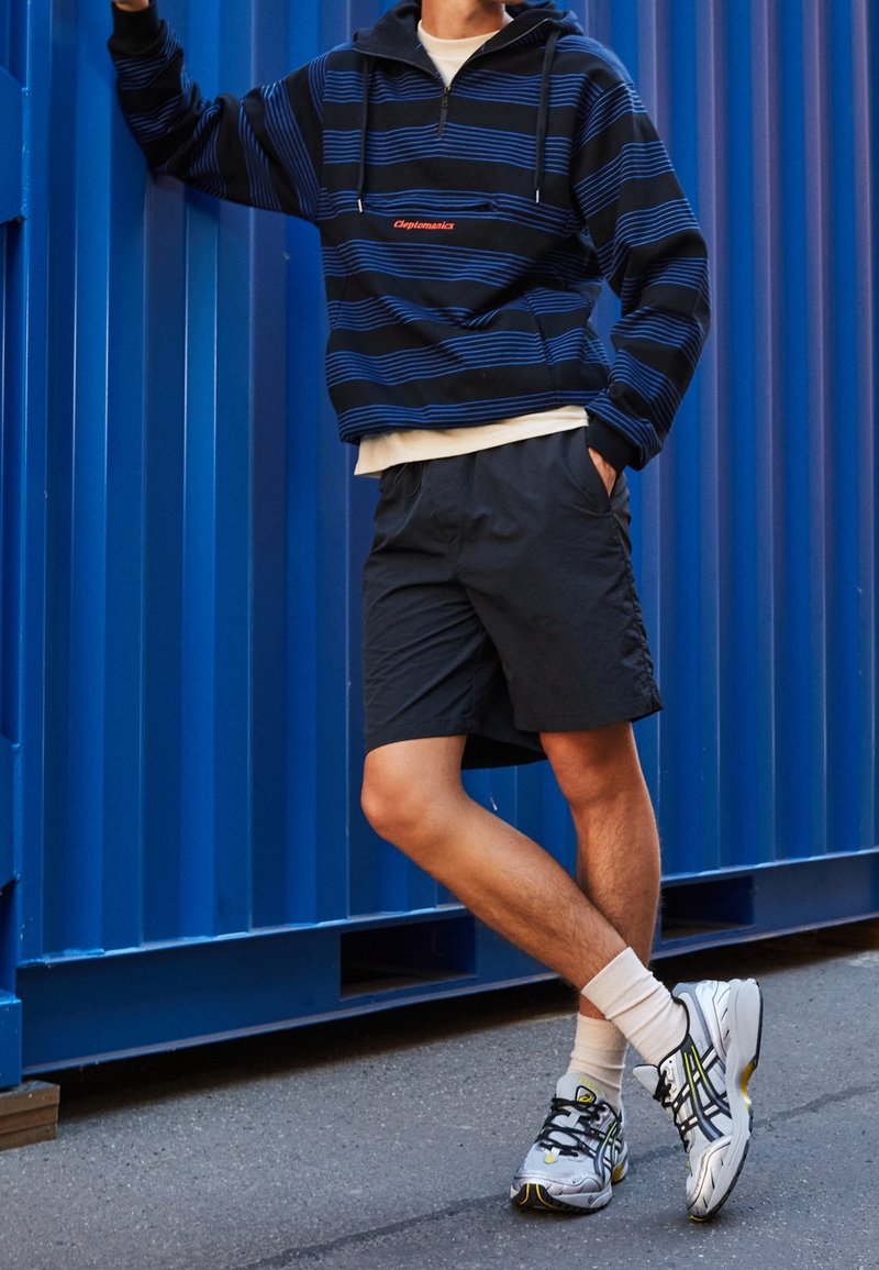 Maglione a righe blu navy con mezza cerniera, pantaloncini sportivi neri e scarpe da corsa argentate con dettagli gialli. Calzini bianchi visibili.