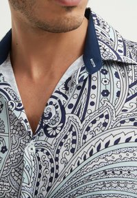 Chemise à manches courtes avec un motif cachemire bleu et blanc, dotée d'un col bleu marine uni et d'une fermeture par bouton. Le tissu semble léger et lisse.