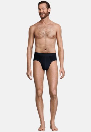 Schwarze Stoffunterhosen mit glatter Textur, die eine enge Passform und einen Bündchen besitzen. Das Modell hat helle Haut und kurze Haare und steht barfuß.