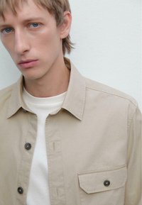 Ung mand med lyst blondt hår og blå øjne bærer en beige skjorte med knapper over en hvid t-shirt foran en ensfarvet hvid baggrund.
