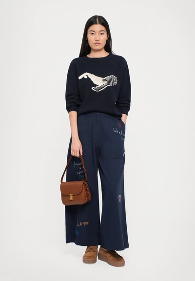 Navy sweater with a cream-colored bird design, paired with wide navy trousers featuring embroidered text. Brown crossbody bag and tan platform shoes.