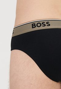 Juodos medvilninės kelnaitės su plačia chaki spalvos juosmens juosta, ant kurios juodais raidėmis parašyta „BOSS“, akcentuota plona balta juostele. Minkšta tekstūra.