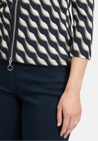 Kurtka zapinana na zamek błyskawiczny z falistym wzorem w kolorze granatowym, białym i zielonym; tekstura prążkowana; rękawy 3/4; zestawiona z ciemnogranatowymi spodniami. Złoty akcent przy zamku.