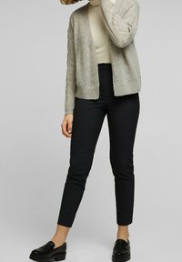 Ljusgrå stickad cardigan med ett texturerat mönster, bärs över en krämfärgad polotröja, i kombination med svarta figurnära byxor och svarta loafers.