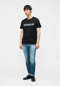 Άνδρας που φοράει μαύρο t-shirt DONdup, ανοιχτόχρωμο μπλε τζιν με μανσέτες και μαύρα μοκασίνια, στέκεται μπροστά σε λευκό φόντο.