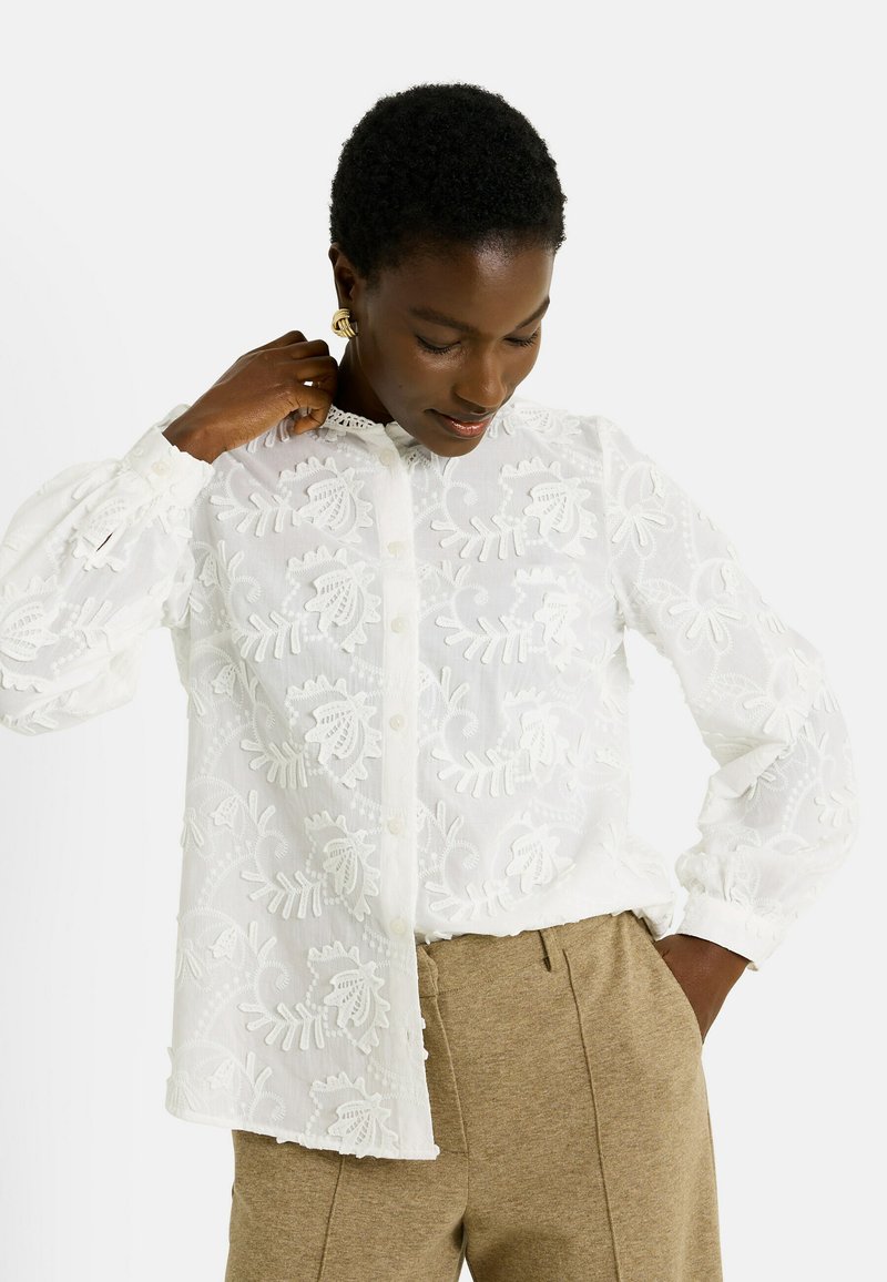 Blusa blanca bordada con patrones florales, botones en la parte delantera y mangas largas. Presenta un delicado cuello y una tela texturizada.