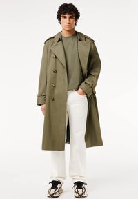 Trench coat verde oliva con design doppiopetto, bottoni dorati e spalline; indossato sopra una maglietta verde e pantaloni bianchi.