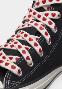 Μαύρο αθλητικό παπούτσι με λευκά κορδόνια που φέρει επαναλαμβανόμενο μοτίβο μικρών κόκκινων καρδιών, περασμένα από μεταλλικά σκρουτζ.