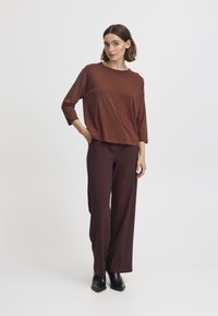 Maglia lunga a maniche lunghe, di colore marrone e taglio largo, realizzata in materiale morbido, abbinata a pantaloni larghi marrone scuro e stivaletti neri alla caviglia.