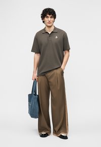 Νεαρός άνδρας με σγουρά μαλλιά, φορώντας ελιά πόλο και φαρδιές καφέ παντελόνες, κρατώντας μια μπλε υφασμάτινη τσάντα χειρός, στέκεται μπροστά σε λευκό φόντο.