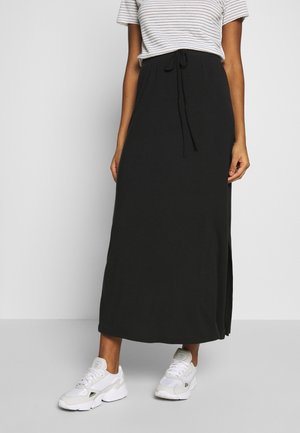 Vrouw draagt een zwarte enkellange rok met een taillekoord, een gestreept T-shirt met korte mouwen en witte sneakers tegen een effen achtergrond.