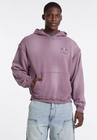 Felpa con cappuccio viola con toppa logo, texture morbida, orlo grezzo e tasca frontale. Abbinate a jeans chiari, rovinati.