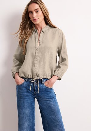 Frau mit langen blonden Haaren, die ein beiges Hemd mit Knopfleiste und Kordelzug in der Taille sowie blaue Jeans trägt, mit den Händen in den Taschen, stehend vor weißem Hintergrund.