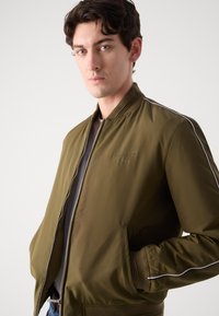 Olive green bomber jacket with a zip front, ribbed collar, and cuffs, featuring white side stripes and an embroidered logo on the chest.