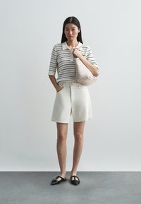 Fehér kötött galléros póló fekete csíkokkal, fehér rövidnadrág, fekete lapos cipő és fonott világos rózsaszín táska. Egyszerű, hétköznapi öltözék.