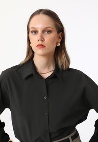 Black button-up shirt with a structured collar and long sleeves, made from a smooth material. Paired with a simple gold necklace.