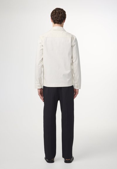 Ljus beige skjorta med lång ärm, knapplås och rak skärning. Matchad med mörka ribbade byxor som visar upp en avslappnad passform.