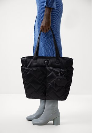 Sac cabas matelassé noir avec deux poches extérieures et une pochette ronde détachable, tenu par une personne portant une robe bleu côtelée et des bottines grises à talons.