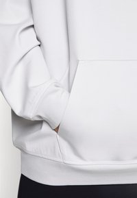 Personne portant un sweat-shirt blanc avec des poignets côtelés, main partiellement à l'intérieur de la poche kangourou à l'avant, pantalons noirs visibles en bas.
