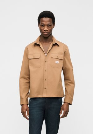 Homme portant une chemise beige boutonnée avec deux poches avant et un jean bleu foncé, debout devant un fond blanc uni.