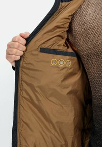 Wasserabweisende Jacke mit glattem, dunklem Außenmaterial und einer kontrastierenden braunen Fütterung. Die Innentasche verfügt über Symbole für Wasser- und Fleckenresistenz.