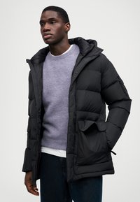 Μαύρο μπουφάν με κουκούλα, με καπιτονέ σχέδιο και τσέπες με φερμουάρ, φορεμένο πάνω από ένα ανοιχτό μωβ πουλόβερ και μπλε τζιν.