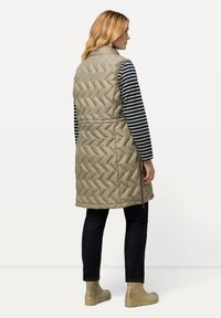 Khaki pikowana kamizelka z wysokim kołnierzem, wzorem w zygzaki, bocznymi kieszeniami, zestawiona z granatową i białą w paski koszulą z długim rękawem oraz ciemnymi dżinsami.