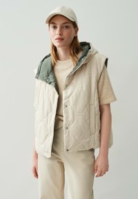 Junge Frau, die eine beigefarbene gesteppte ärmellose Jacke mit einer grünen Kapuze, ein beiges Strickoberteil, eine Hose und eine hellfarbige Mütze trägt und vor einem weißen Hintergrund steht.